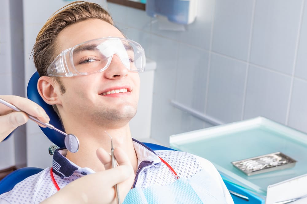 A man wearing protective glasses sits in a dental chair, smiling as the dentist prepares dental tools. - Dental Implants in Palm Springs