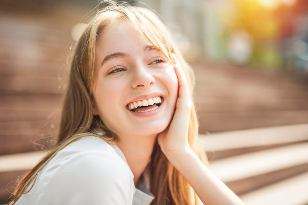 Young Woman Smiling Outdoors - Cosmetic Dentistry A cheerful young woman with blonde hair smiles brightly while resting her hand on her face in natural sunlight. - Cosmetic Dentistry