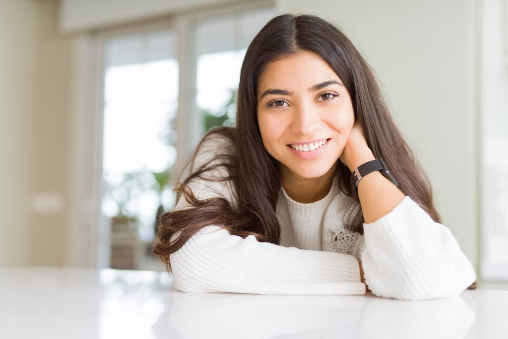 Smiling young woman with straight white teeth, resting her arms on a table. - Veneers A cheerful young woman with long dark hair and a bright, radiant smile sits at a white table in a sunlit room. - Veneers