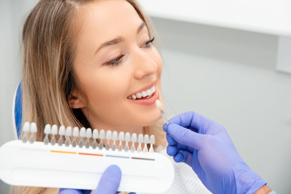A smiling woman at the dentist undergoes a dental consultation. The dentist, wearing gloves, holds up a tooth shade guide for a natural and bright smile. - Veneers in Palm Springs