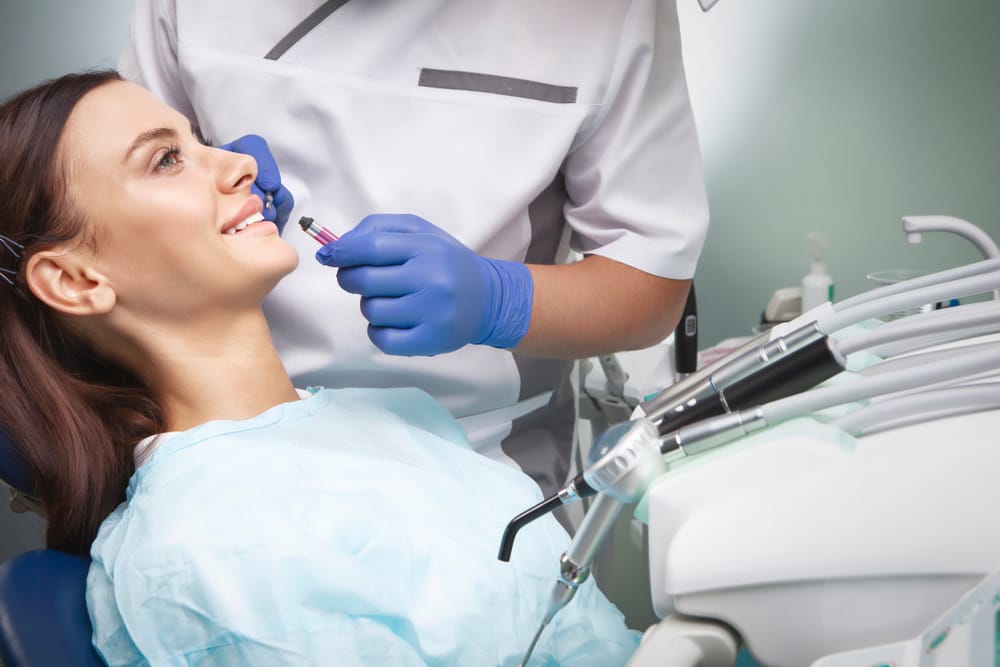 A woman lies in a dental chair smiling while a dentist wearing gloves uses dental instruments to perform a treatment. - Dental Implants