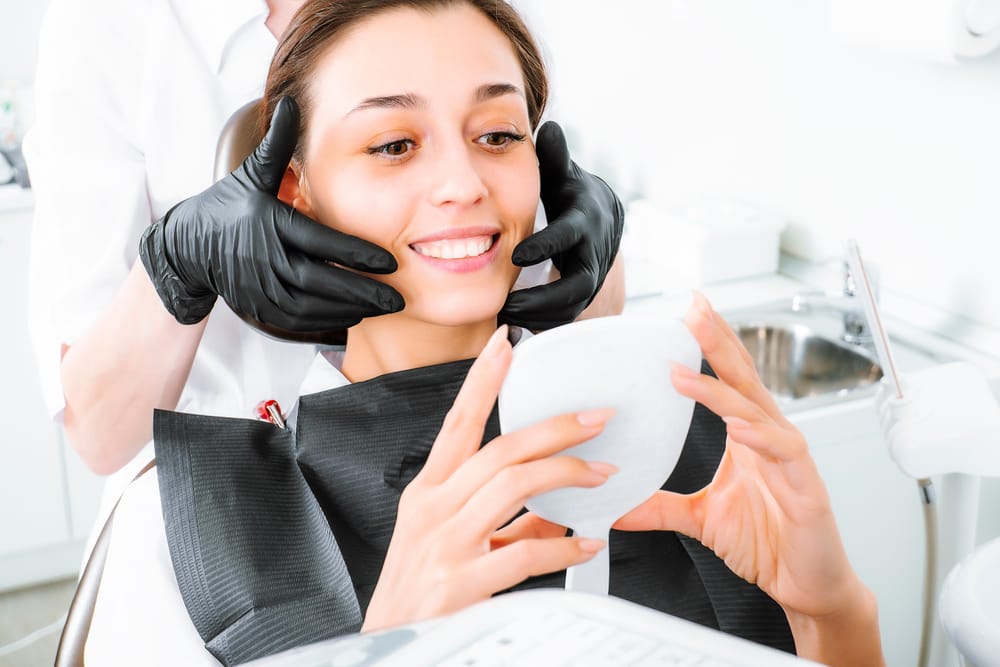 Patient Smiling at Dental Results - Prosthodontics Dentistry A woman holds a mirror and smiles while the dentist adjusts her face and jaw to check her dental results. - Prosthodontics Dentistry
