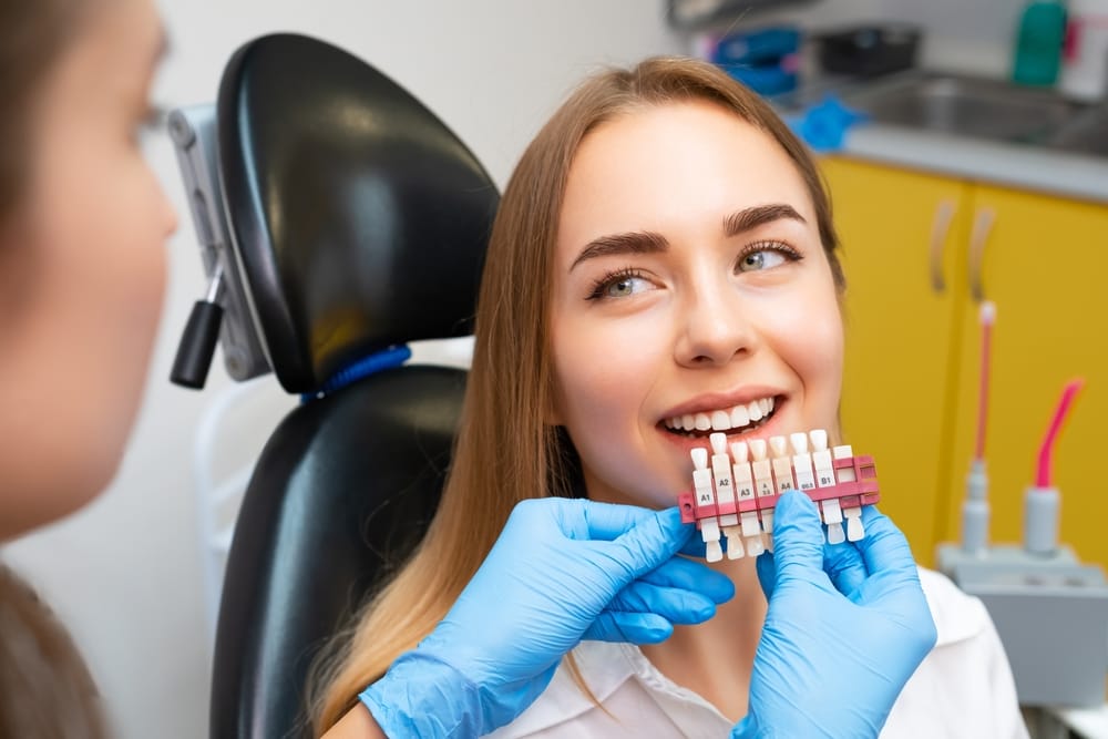 Patient Choosing Tooth Shade at Dentist - Composite Veneers A young woman sits in a dental chair smiling while a dentist wearing blue gloves holds a tooth shade guide to match the color of her teeth for cosmetic or restorative treatment. - Composite Veneers