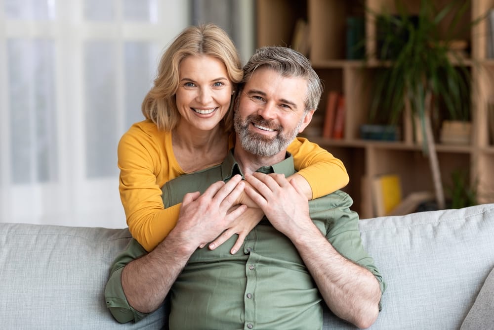 A cheerful couple sits on a couch, smiling warmly as the woman embraces the man from behind. - Dentist in Palm Springs