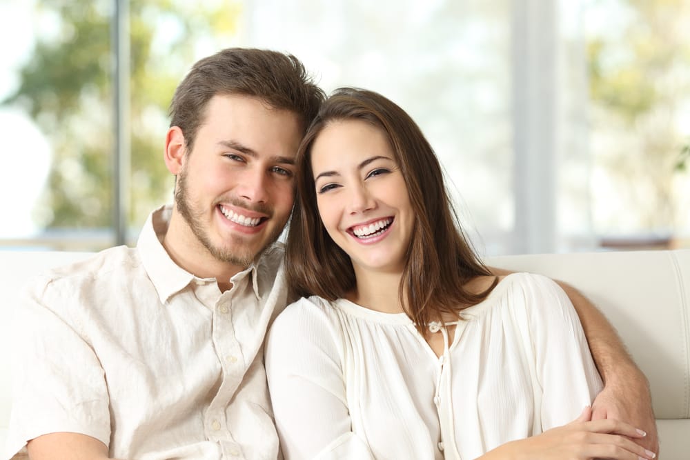 A happy young couple sits close together on a couch, smiling brightly at the camera. - Dentist in Palm Springs