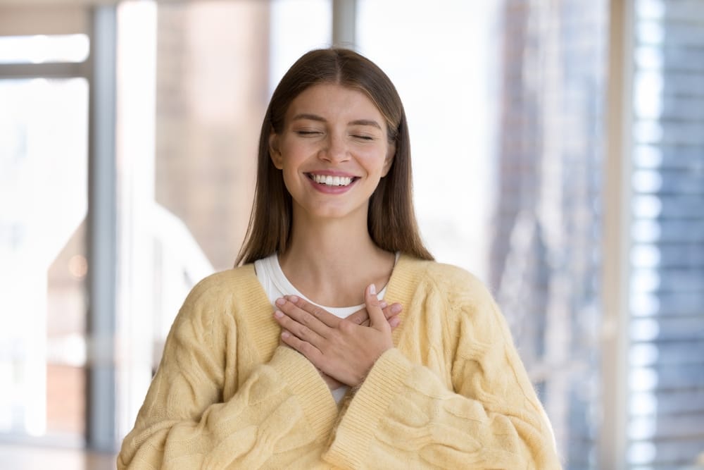 A young woman smiles with closed eyes, holding her hands over her chest while wearing a cozy yellow sweater indoors. - Diagnostic and Preventative
