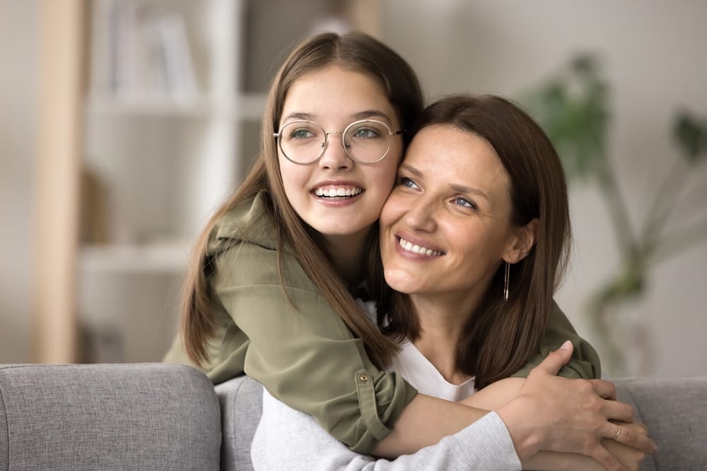 Mother and Daughter Hugging - Dentist in West Palm Beach A smiling daughter wearing glasses and braces hugs her mother lovingly as they both look happy together. - Dentist in West Palm Beach
