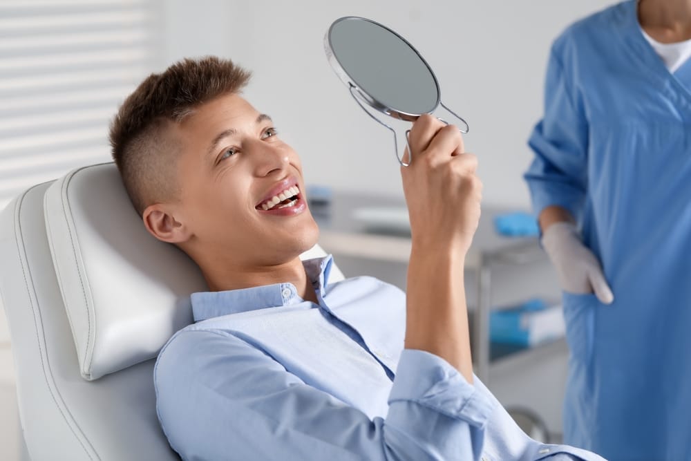 A young man in a dental chair smiles while looking at his teeth in a hand mirror. - Dental Crowns in Palm Springs