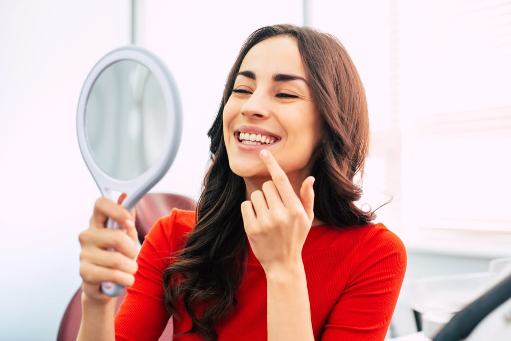 Woman Smiling in Dental Office with Mirror - Composite Veneers A woman in a red shirt sits in a dental chair, smiling while holding a mirror and pointing at her teeth, admiring her smile. - Composite Veneers