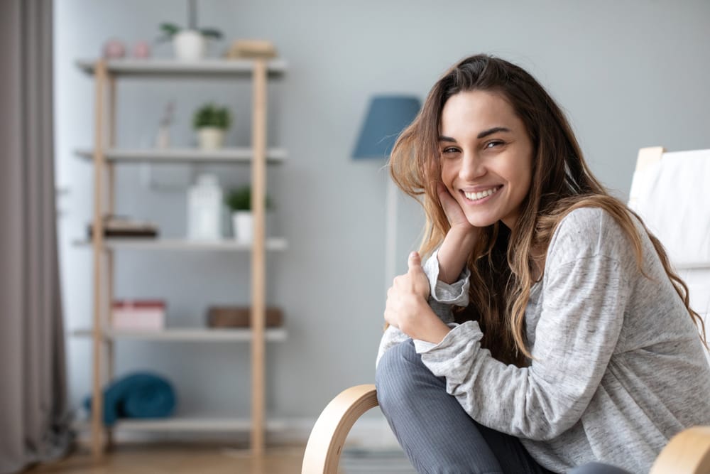Smiling young woman sitting on a chair. - Composite Veneers A cheerful young woman with long hair sits comfortably in a chair, smiling warmly in a minimalist living room. - Composite Veneers