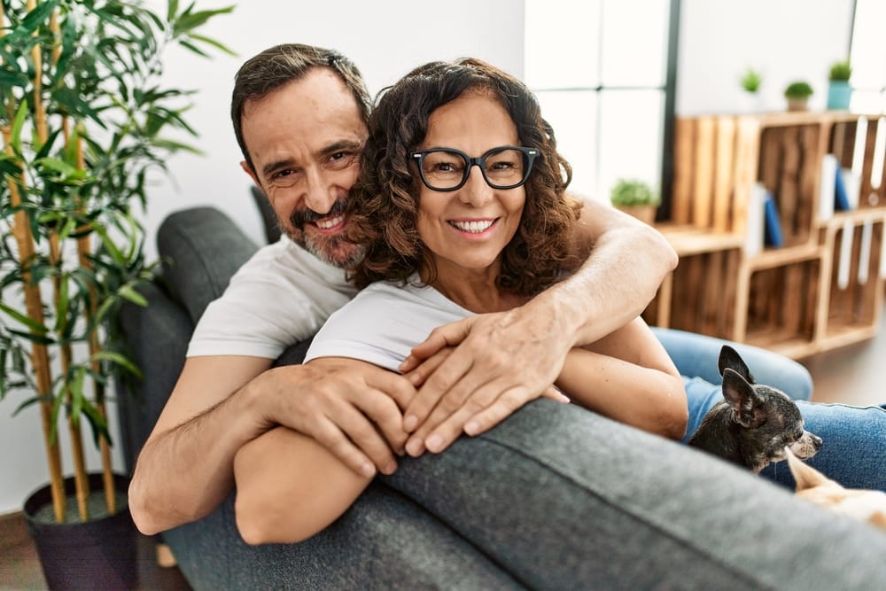 Happy Couple Relaxing at Home - Dentist in West Palm Beach A smiling couple sits on a couch, embracing each other warmly, with a small dog resting beside them. - Dentist in West Palm Beach