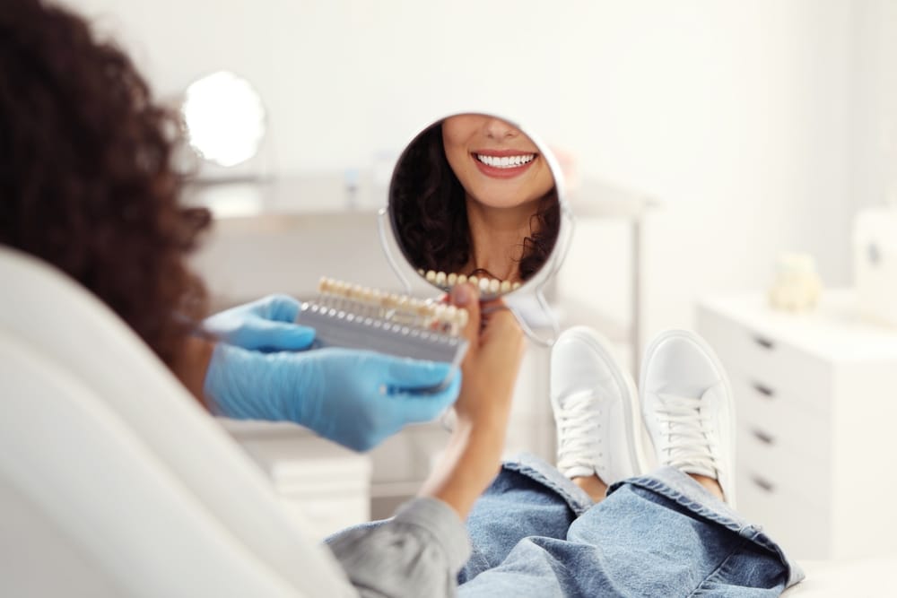 Patient Smiling in Dental Mirror - Cosmetic Dentistry A dental patient looks into a hand mirror, smiling while the dentist holds a tooth shade guide for comparison. - Cosmetic Dentistry