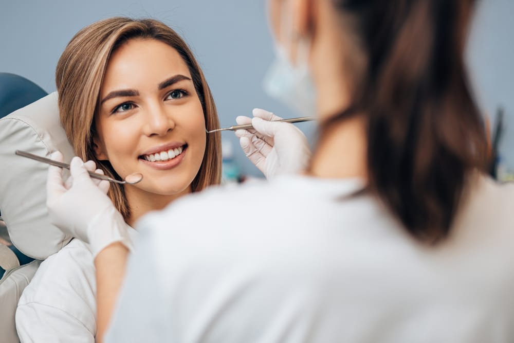 Woman Smiling During Dental Checkup - Oral Surgery A young woman sits in a dental chair, smiling as a dentist examines her teeth with dental instruments. - Oral Surgery