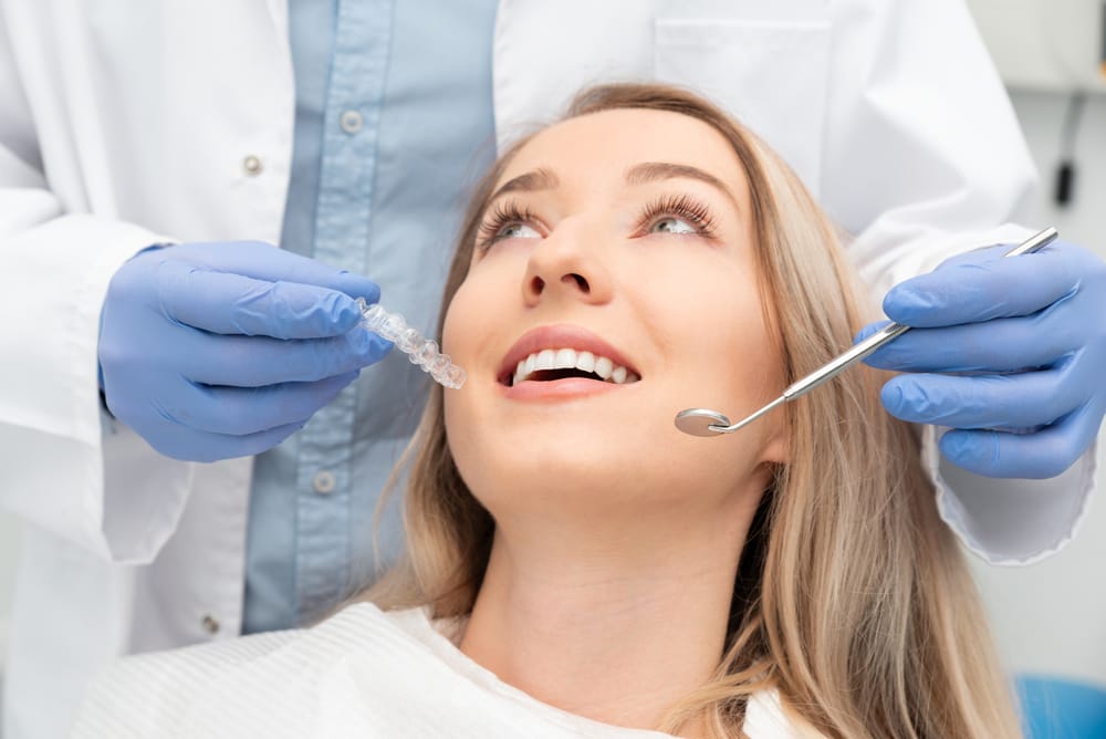 A woman in a dental chair smiles while a dentist holds a clear aligner and dental tool near her mouth. - Dental Bridges in Palm Springs