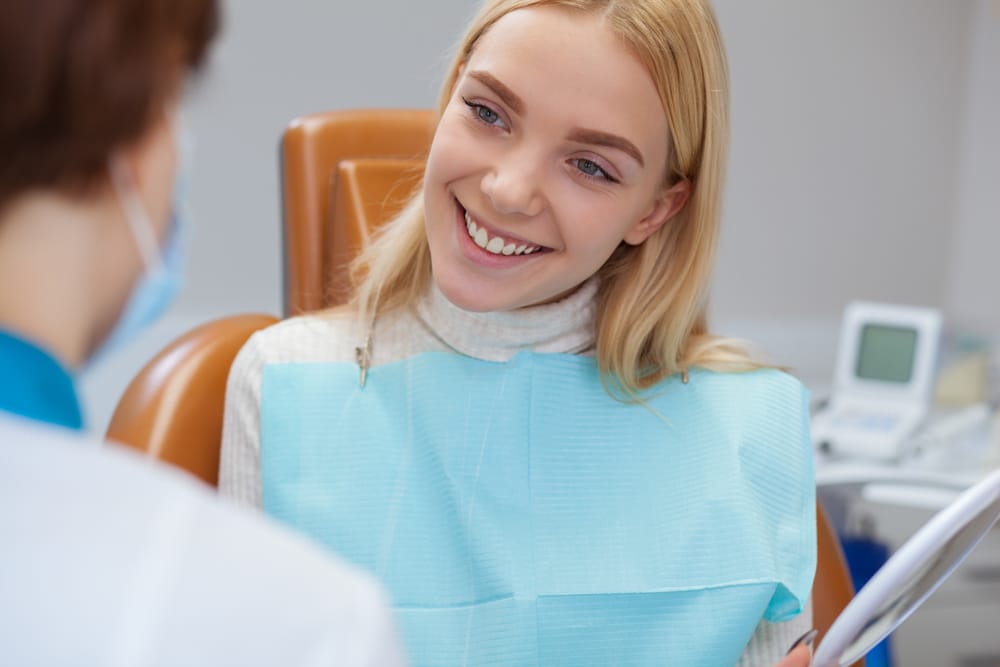 A young woman wearing a dental bib smiles while speaking with a dentist during her appointment. - Diagnostic and Preventative