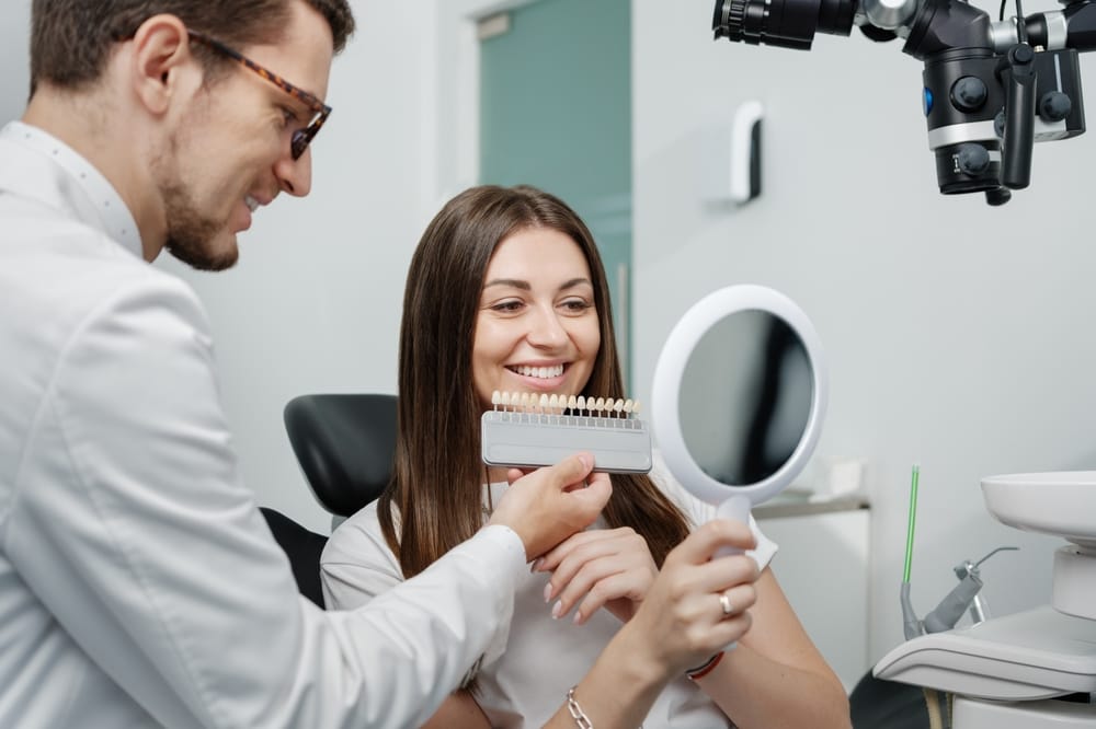 A smiling woman holds a mirror while a dentist shows her a tooth shade guide to select the best color for dental treatment. - Veneers in West Palm Beach