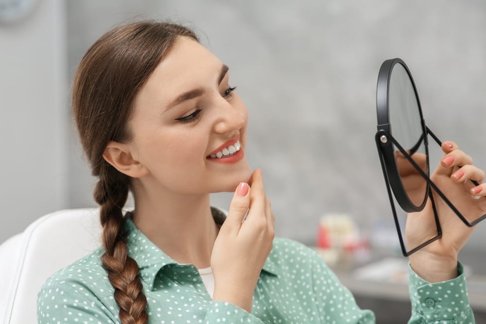 A young woman with a braid smiles while examining her teeth in a handheld mirror at the dentist’s office. She looks pleased with her bright, healthy smile. - Veneers in Palm Springs