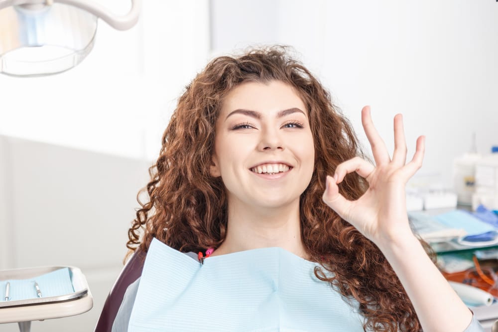 A young woman with curly hair sits in a dental chair wearing a bib, smiling and making an "OK" hand gesture, showing satisfaction with her dental visit. - Smile Design