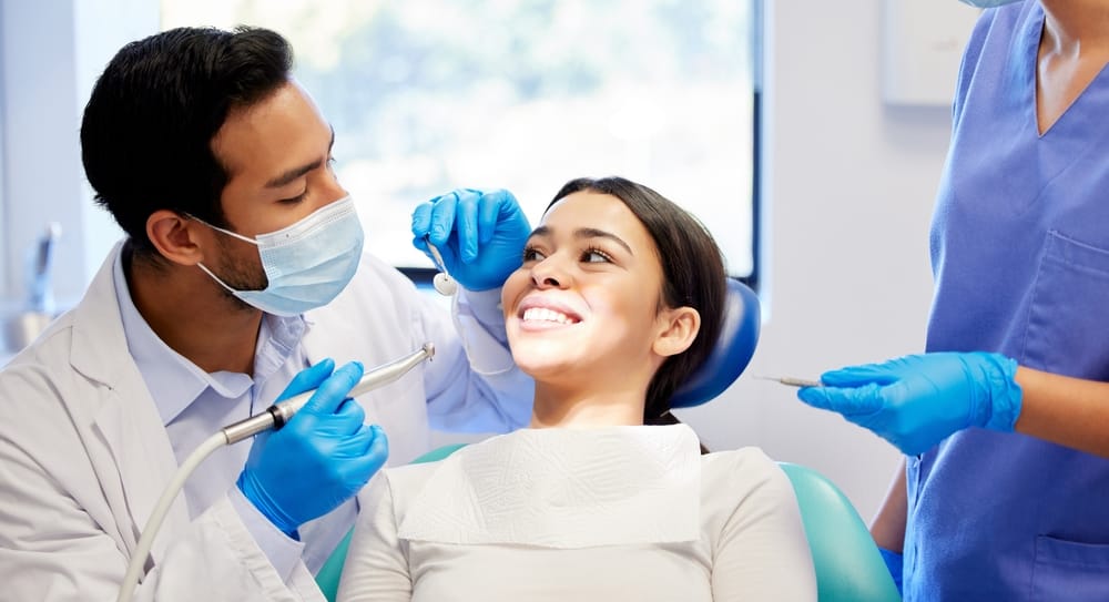 A woman sits in a dental chair smiling while a dentist in a mask and gloves examines her teeth with dental instruments, assisted by a dental nurse. - Oral Surgery
