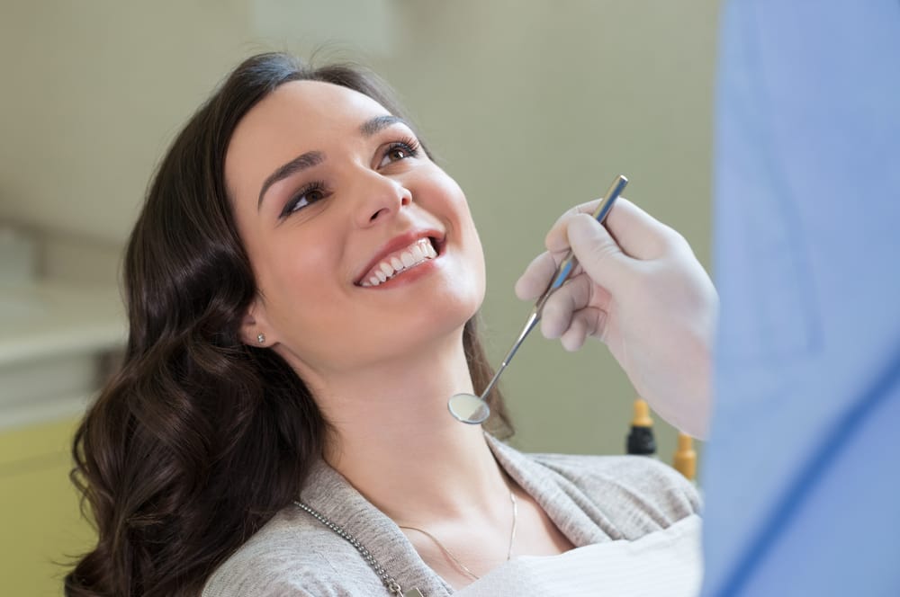 A woman sits in a dental chair smiling while a dentist examines her teeth with a dental mirror. - Orthodontics