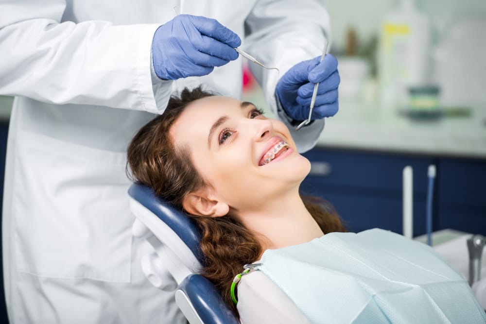 A smiling woman with braces sits in a dental chair while the dentist prepares dental tools for her checkup. - Endodontics