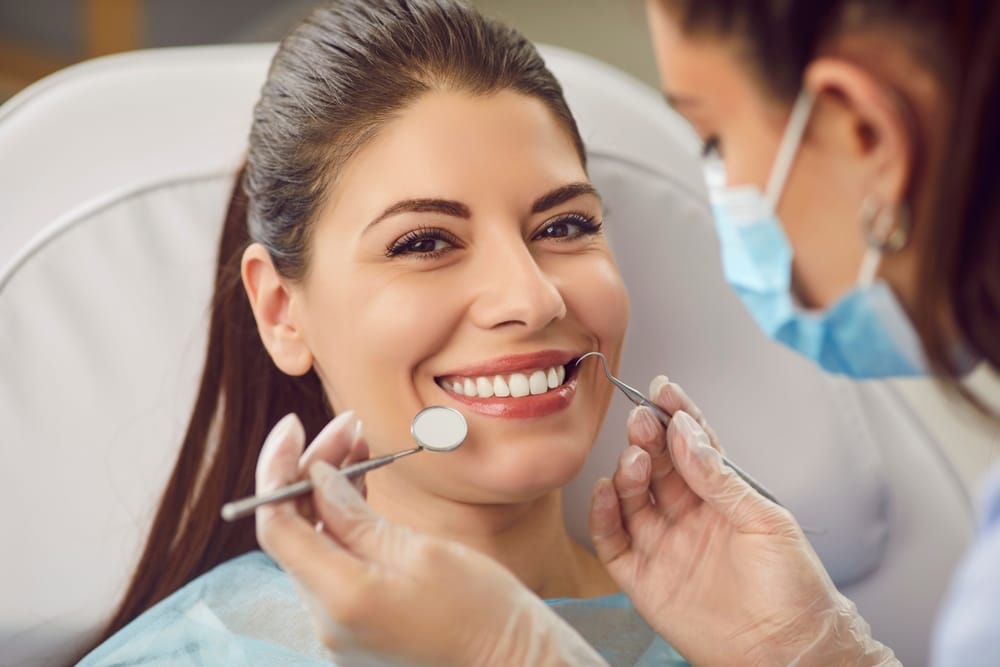 A woman in a dental chair smiles while a dentist examines her teeth with dental instruments. - Endodontics