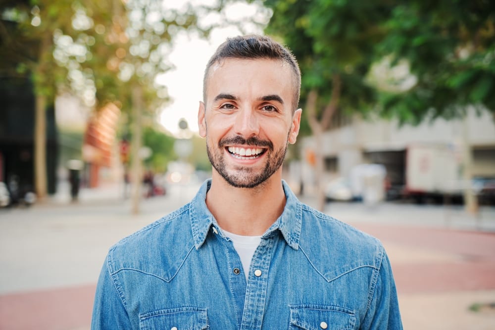 Smiling Man Outdoors - Veneers A cheerful man in a denim shirt smiles brightly while standing on a city street with trees and buildings in the background. - Veneers