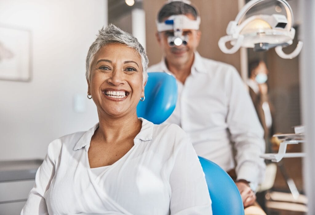 Smiling Woman at the Dentist - Services A woman with short gray hair smiles confidently while sitting in a dental chair. Behind her, a dentist wearing magnifying glasses is preparing for the checkup. - Services