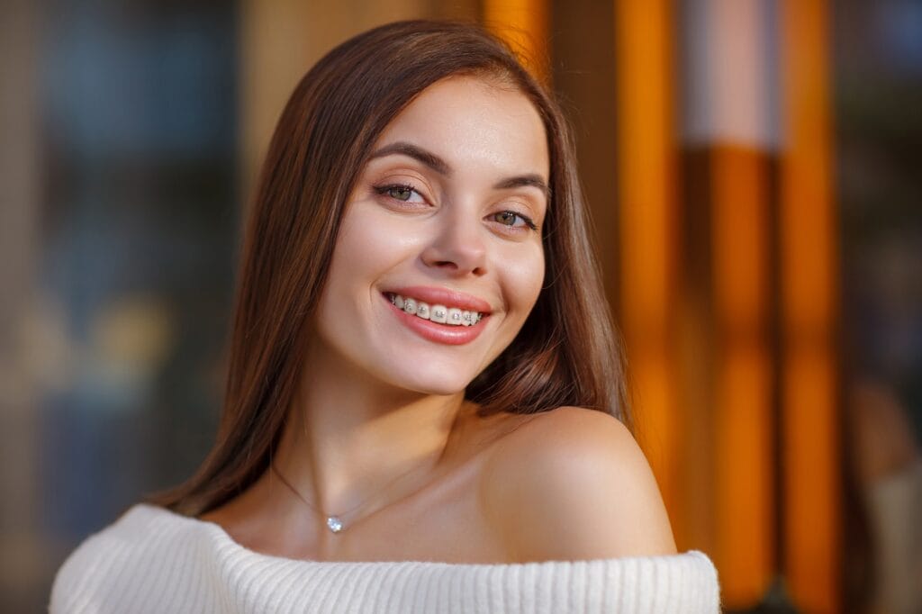 Smiling Young Woman with Braces - Services A young woman with long brown hair smiles brightly, showing her braces. She is wearing an off-shoulder white sweater and a delicate necklace, with a warm blurred background that enhances her cheerful expression. - Services