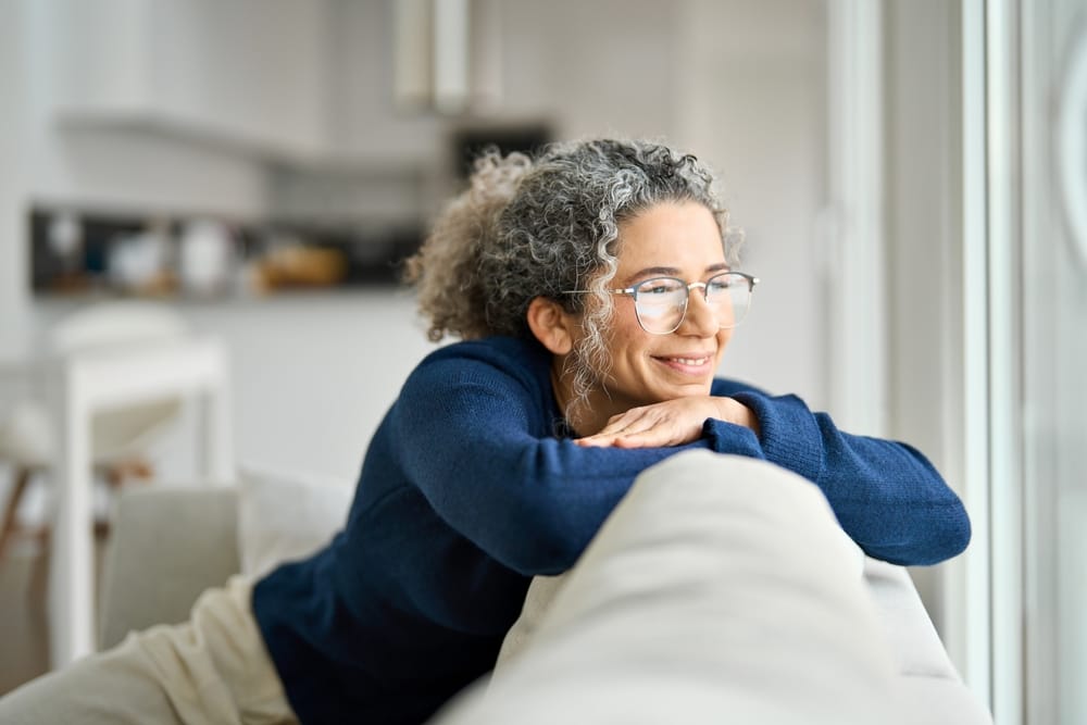Smiling Woman Looking Out Window - Dentist in West Palm Beach A woman with curly gray hair and glasses smiles warmly while leaning on the back of a couch, gazing outside. - Dentist West Palm Beach