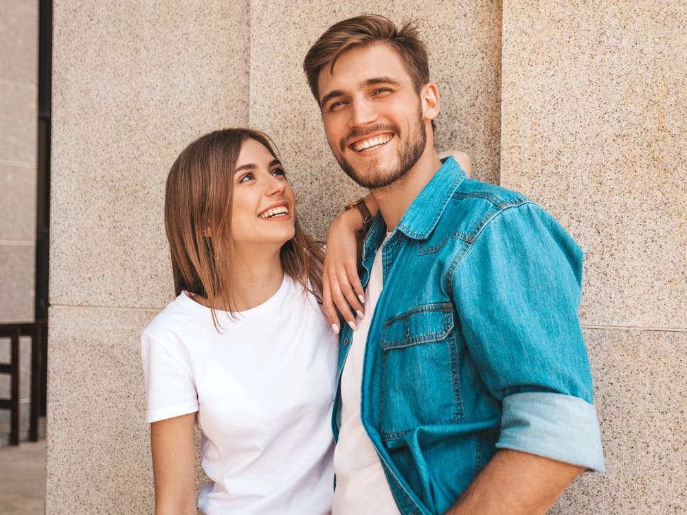A cheerful young couple stands by a wall, smiling warmly. The woman in a white t-shirt looks up at the man in a denim jacket, capturing a moment of happiness and closeness. - Smile Design