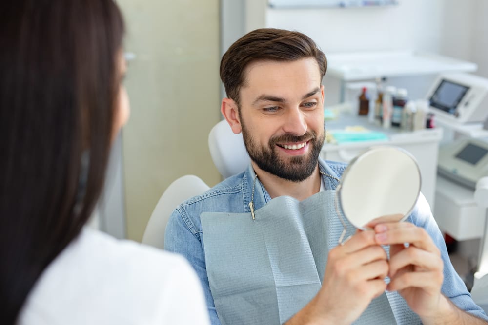 Man at the dentist checking his teeth - Dental Implants in West Palm Beach A man sits in a dental chair, happily looking at his reflection in a small mirror after treatment. - Dental Implants in West Palm Beach