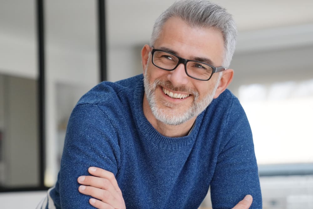 A man with gray hair, beard, and glasses smiles warmly while wearing a blue sweater indoors. - Dental Crowns in Palm Springs