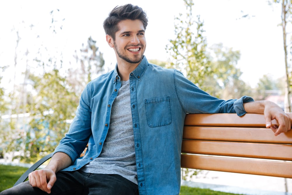 A young man in a denim shirt sits on a wooden bench outdoors, smiling and enjoying a sunny day surrounded by trees. - Dental Implants